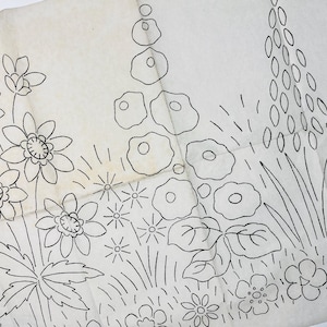 May include: A white paper embroidery transfer pattern featuring black line art illustrations of various garden flowers, including daisies, foxgloves, and leafy foliage, arranged in a botanical composition for needlework projects.