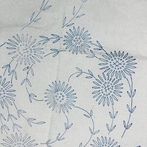 May include: A white fabric sheet featuring a blue ink transfer pattern of multiple daisy-like flowers with long, thin stems and small leaves. The floral design is arranged in a curved, flowing composition across the surface of the material.