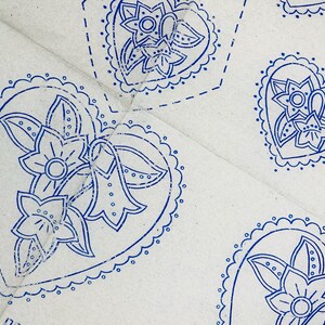 Puede incluir: Una superficie de tela gris claro con un patrón repetitivo estampado en tinta azul. Cada motivo consiste en una forma de corazón con borde festoneado, que contiene un diseño floral central con tres flores estilizadas y hojas. El texto SPECIALLY es visible.