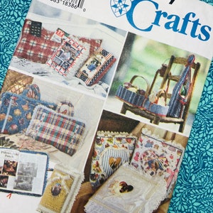 Op de afbeelding: Simplicity Crafts naaipatroon met diverse knutselprojecten, waaronder kussens en decoratieve items. Het patroon bevat afbeeldingen van voltooide projecten met gedetailleerde ontwerpen en versieringen. De achtergrond is een turquoise bloemenpatroon.