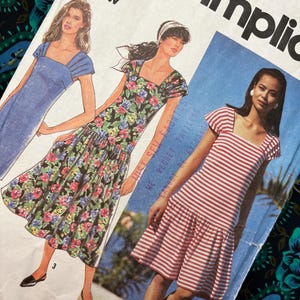 Könnte beinhalten: Ein Simplicity-Schnittmusterumschlag mit Illustrationen von drei Kleidern mit eckigem Ausschnitt und kurzen Ärmeln. Die Designs umfassen ein einfarbig blaues Kleid, ein Kleid mit schwarzem Blumenmuster und ein rot-weiß gestreiftes Kleid mit tiefer Taille.