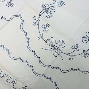 Può includere: Tessuto bianco con ricami floreali blu e bordi smerlati. Il ricamo presenta trifogli stilizzati a quattro foglie e delicate viti. Il testo "TRANSFER for TOW" è visibile in basso a sinistra.