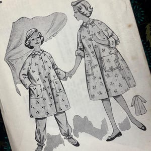 Op de afbeelding: Een vintage naaipatroon-envelop met een zwart-witillustratie van twee figuren in jassen met bloemenprint, voorzien van kragen en zakken. Eén figuur draagt een broek met omslag, de andere een jurk. Tekst: Turnings are allowed on this pattern.