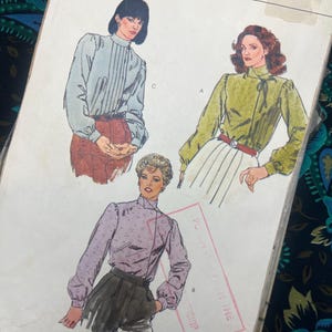 Op de afbeelding: Een Vogue naaipatroon-envelop met illustraties van drie blouses met lange mouwen en een hoge halslijn. De ontwerpen omvatten een lichtblauwe geplooide blouse, een groene blouse met een strikceintuur en een lila blouse met patroon en zijsluiting.