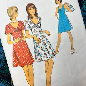 Pode incluir: Um envelope de molde de costura com ilustrações de três vestidos. Um é vermelho com bolinhas brancas, um é branco com estampa floral azul e vermelha e um é azul sólido. Cada vestido tem um decote redondo e mangas curtas ou sem mangas.
