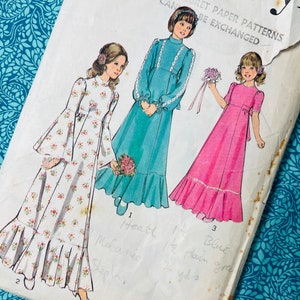 May include: A vintage sewing pattern envelope for size 10, breast 73 cm, featuring illustrations of three floor-length dresses. The designs include a floral print dress with bell sleeves, a teal dress with lace trim, and a pink dress with short sleeves.