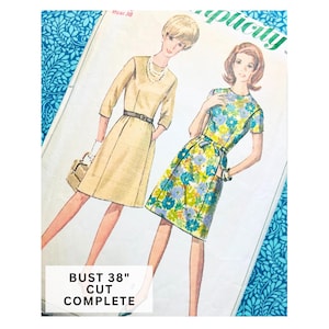 May include: A Simplicity sewing pattern envelope featuring illustrations of two dresses. One is a solid tan dress with a belt and three-quarter sleeves, the other is a floral print dress with short sleeves. Text reads BUST 38" CUT COMPLETE.