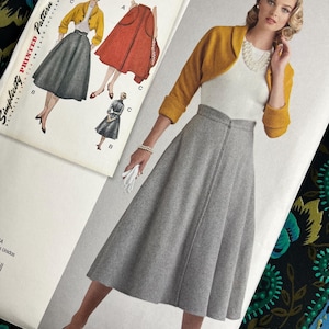 Op de afbeelding: Een vrouw draagt een grijze rok en een mosterdgele bolero. Een vintage Simplicity patroon is zichtbaar, met illustraties van de outfit. Het patroonpakket is wit met zwarte tekst. De achtergrond heeft een bloemenpatroon.