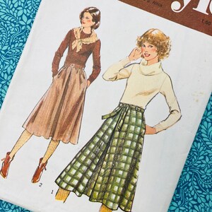 May include: A sewing pattern envelope labelled Style featuring illustrations of two outfits. One shows a brown long-sleeve top with a scarf and a pleated skirt. The other shows a cream turtleneck sweater with a green and white plaid skirt with a belt.