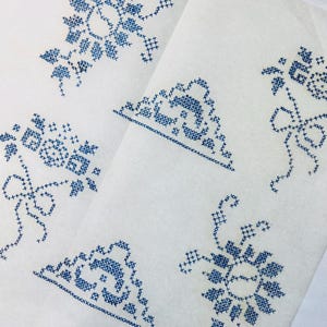 May include: Close-up of several pieces of white fabric with blue cross-stitch embroidery. The embroidery features floral and geometric designs, including flowers, leaves, and bows. The fabric pieces are arranged at different angles.