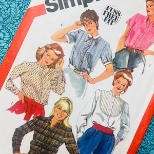 May include: A Simplicity sewing pattern envelope featuring illustrations of five different blouse styles. The designs include a plaid shirt, a floral print blouse, a striped shirt with a collar, a pink button-down, and a white blouse with a pleated bib.