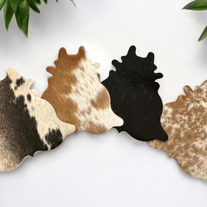 May include: Four cowhide coasters in different patterns. Two are brown and white, one is black, and one is brown and white with a more mottled pattern.