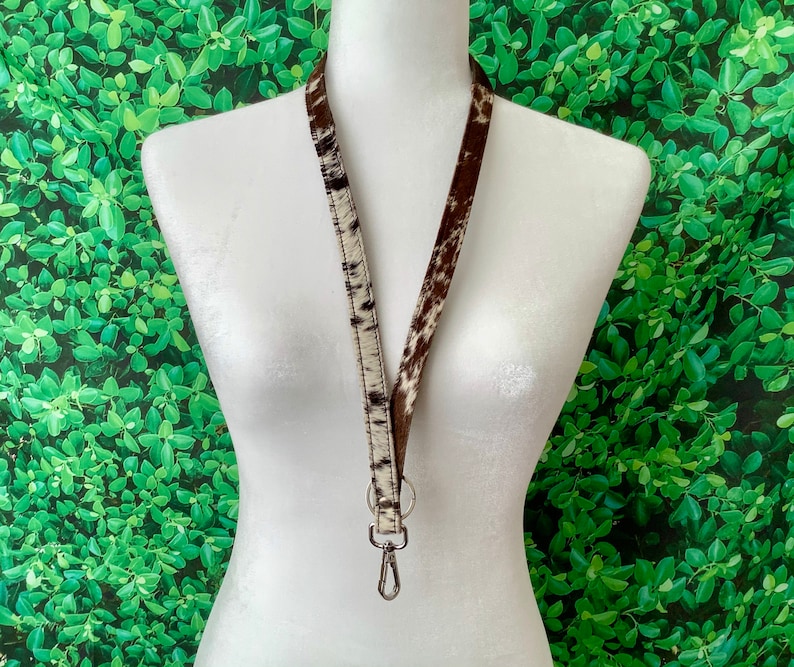 Op de afbeelding: Een wit en bruin koeienhuid lanyard met een zilveren draaibare clip. Het lanyard hangt aan een wit mannequin.