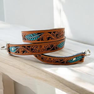 May include: Two brown leather straps with intricate floral and leaf patterns in black and turquoise. The straps have silver-colored metal clasps and are likely for a bag or purse. The leather has a distressed look.