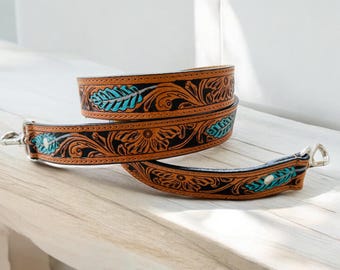 May include: Two brown leather straps with detailed floral and leaf designs in black and turquoise. The straps have silver metal clasps, possibly for a bag. The leather has a worn appearance. The straps are approximately 100 cm long.