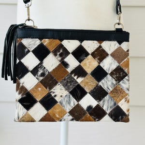 Puede incluir: Bolso bandolera rectangular con diseño patchwork. El bolso tiene una parte superior y una correa de cuero negro, un cierre de cremallera y una borla negra. El patchwork está hecho de cuadrados en negro, blanco y tonos marrones.