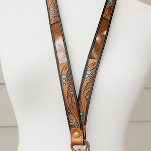 May include: A brown leather lanyard with a cowhide pattern and turquoise accents. The lanyard features a decorative floral design and a metal clasp. The design includes a mix of brown, white, and turquoise colors.