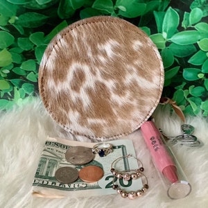 May include: A round, brown and white cowhide coin purse with a zipper closure. The purse is open and contains US currency, rings, and a pink lip balm tube labeled "ROSA".