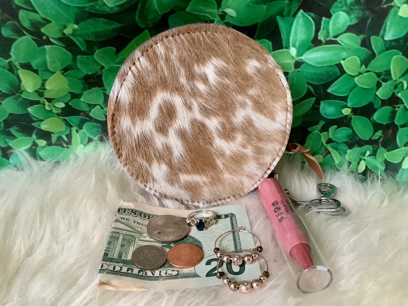 May include: A round, brown and white cowhide coin purse with a zipper closure. The purse is open and contains US currency, rings, and a pink lip balm tube labeled "ROSA".
