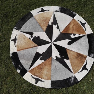 May include: A round cowhide rug with a starburst design in black, white, and brown. The rug is made of different pieces of cowhide sewn together.