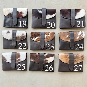 May include: Nine leather clutch bags with cowhide flap closures. Each bag has a dark brown leather body and a flap made of cowhide in various patterns of brown, white, and black. Numbers 19 to 27 are printed on the bags.
