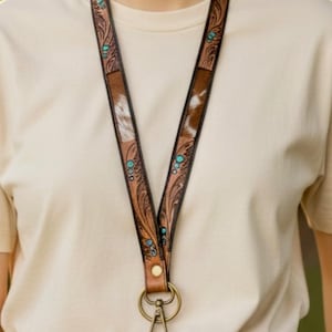 May include: A brown leather lanyard with a decorative floral pattern and turquoise accents. The lanyard features a section of cowhide and a metal clasp. The design is visible against a neutral background.