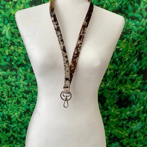 Op de afbeelding: Een bruin en wit koeienhuid lanyard met een zilveren sleutelhanger. Het lanyard hangt aan een wit mannequin.