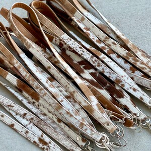 Op de afbeelding: Een groep bruine en witte koeienhuid lederen sleutelhangers met zilveren sleutelringen. De sleutelhangers zijn allemaal even lang en hebben een vergelijkbaar patroon.