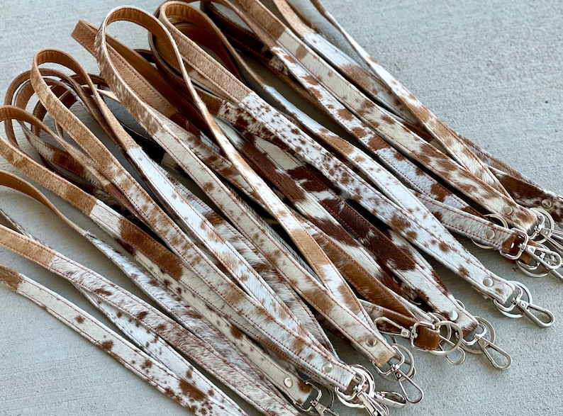 Op de afbeelding: Een groep bruine en witte koeienhuid lederen sleutelhangers met zilveren sleutelringen. De sleutelhangers zijn allemaal even lang en hebben een vergelijkbaar patroon.