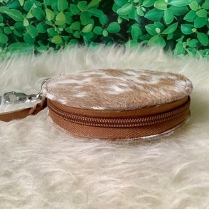May include: A round, brown and white cowhide coin purse with a zipper closure. The purse has a silver key ring attached.