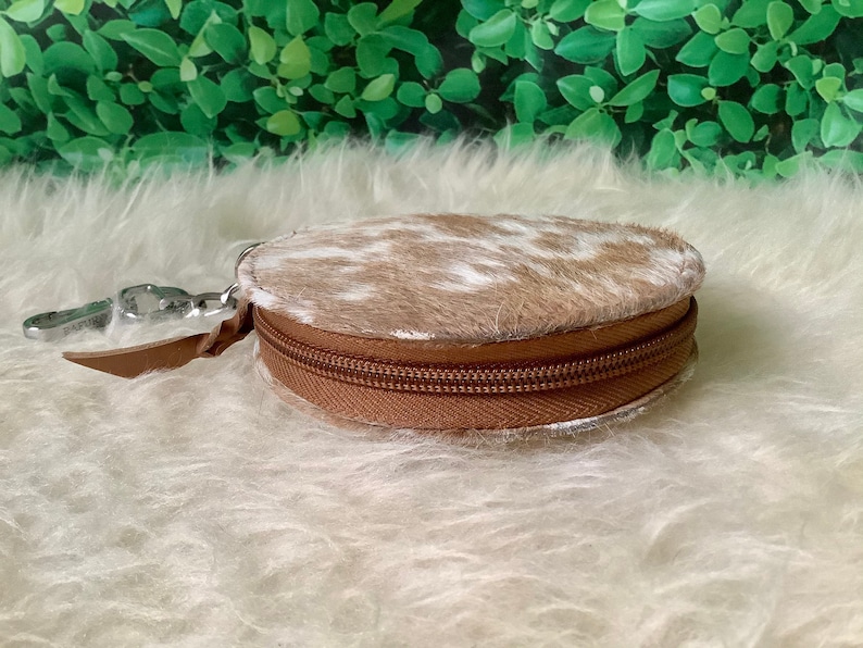 May include: A round, brown and white cowhide coin purse with a zipper closure. The purse has a silver key ring attached.