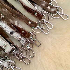 Op de afbeelding: Een groep bruine en witte koeienhuid sleutelhangers met zilveren sleutelringen en sluitingen.