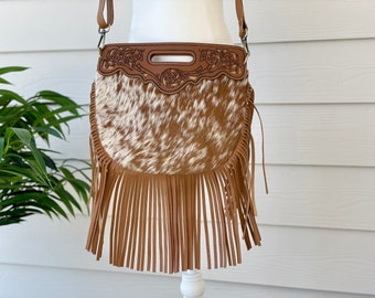 May include: A brown leather crossbody bag with a fringe detail. The bag is made of cowhide and has a floral design on the front flap.