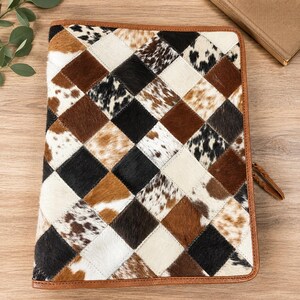 May include: A leather portfolio with a patchwork design. The cover features a diamond pattern of brown, black, and white cowhide. The edges are trimmed with brown leather, and a leather feather charm hangs from the side. The portfolio is designed for documents and papers.