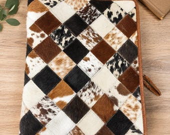 Puede incluir: Un portafolios de cuero con un diseño de patchwork. La cubierta presenta un patrón de diamantes de piel de vaca marrón, negra y blanca. Los bordes están ribeteados con cuero marrón y un adorno de pluma de cuero cuelga del lateral.