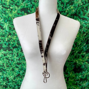 Op de afbeelding: Een zwart-witte koeienhuid lanyard met een zilveren draaibare clip. De lanyard hangt aan een wit mannequin tegen een groene, bladerrijke achtergrond.