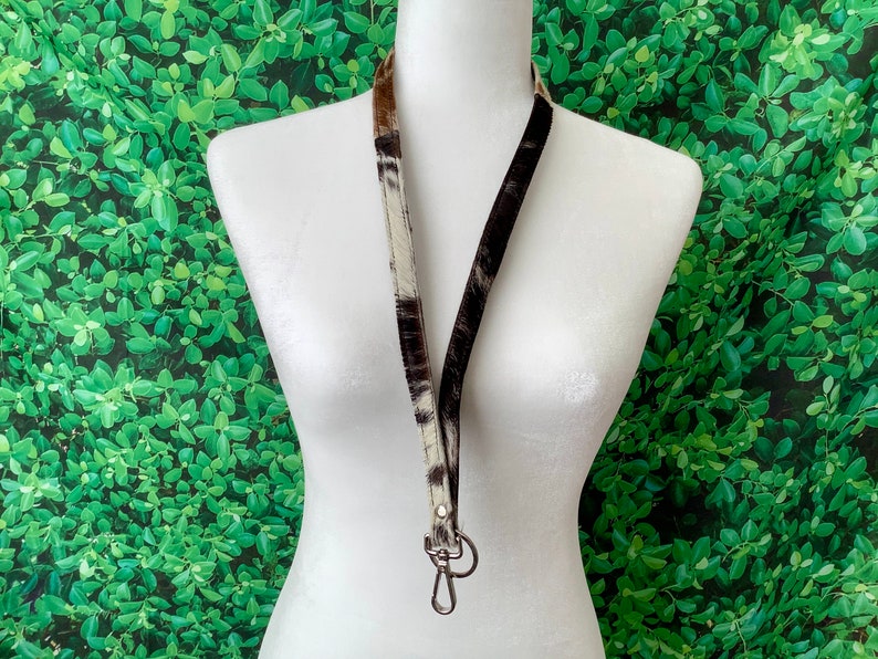 Op de afbeelding: Een zwart-witte koeienhuid lanyard met een zilveren draaibare clip. De lanyard hangt aan een wit mannequin tegen een groene, bladerrijke achtergrond.