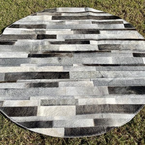 May include: A round, gray and black cowhide rug with a patchwork design. The rug is made of strips of cowhide sewn together in a horizontal pattern.