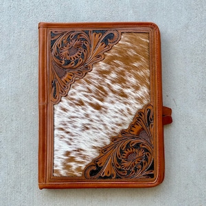 May include: A brown leather folder with a tooled design and a white and brown cowhide pattern. The folder has a flap closure and a strap.