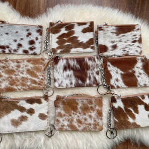 May include: Eight brown and white cowhide leather zippered coin purses with silver keychains. The purses are arranged in two rows of four.