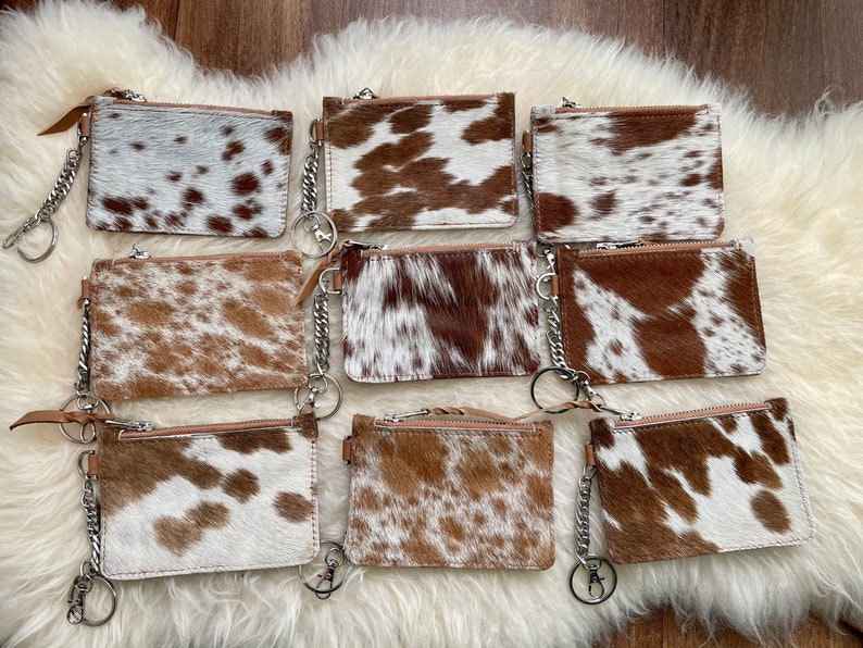 May include: Eight brown and white cowhide leather zippered coin purses with silver keychains. The purses are arranged in two rows of four.