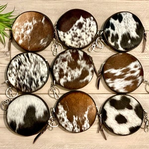May include: Eight round cowhide coin purses with zipper closures and keychains. The purses are in various brown and white patterns.