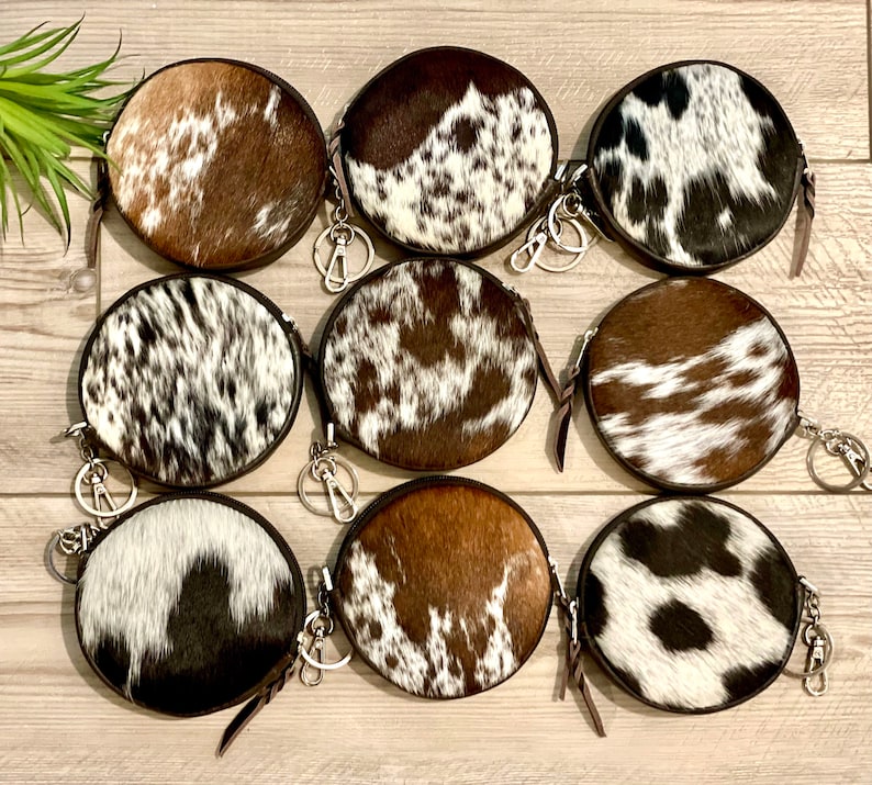 May include: Eight round cowhide coin purses with zipper closures and keychains. The purses are in various brown and white patterns.