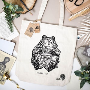 May include: A white tote bag featuring a black and white illustration of a toad. The toad is sitting with its arms crossed, and the text 'Chubby Toad' is printed below it. The text 'Harriet Rose Art' is printed in the bottom right corner of the bag.