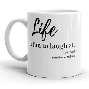 Puede incluir: Taza de cerámica blanca con una inscripción negra que dice "Life is fun to laugh at." La inscripción va seguida del nombre "Kevin Baugh" y el título "Presidente de Molossia."