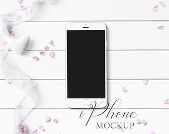 iPhone Mockup Styled with white ribbon, pink flowers, and white heart pillow on a white wood background. Showcase your creations with ease!