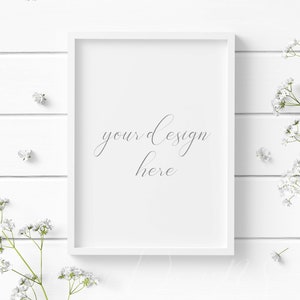 May include: A white rectangular picture frame with the text "your design here" in a script font. The frame is surrounded by white flowers and greenery, set against a white wooden background.