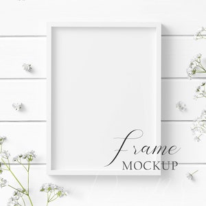 May include: A white rectangular frame mockup with the text "frame MOCKUP" in a script font. The frame is on a white wooden surface, adorned with delicate white flowers and sprigs, creating a clean and elegant aesthetic.