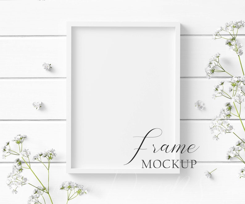 May include: A white rectangular frame mockup with the text "frame MOCKUP" in a script font. The frame is on a white wooden surface, adorned with delicate white flowers and sprigs, creating a clean and elegant aesthetic.