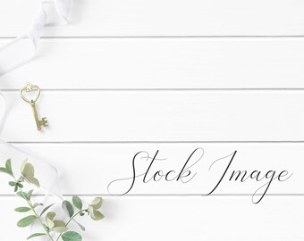 Feminine Flat Lay Styled with greenery, white ribbon, and gold key on a white wood background.